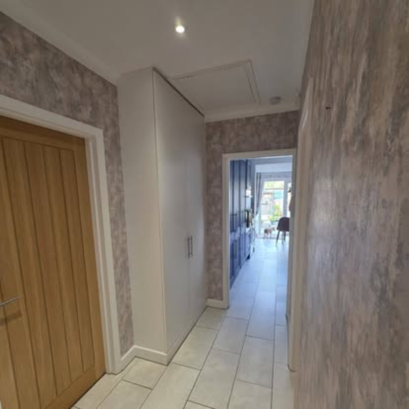 A hallway in grey paper with an oak door on the left and tall white cupboards, looking through an open doorway into a kitchen